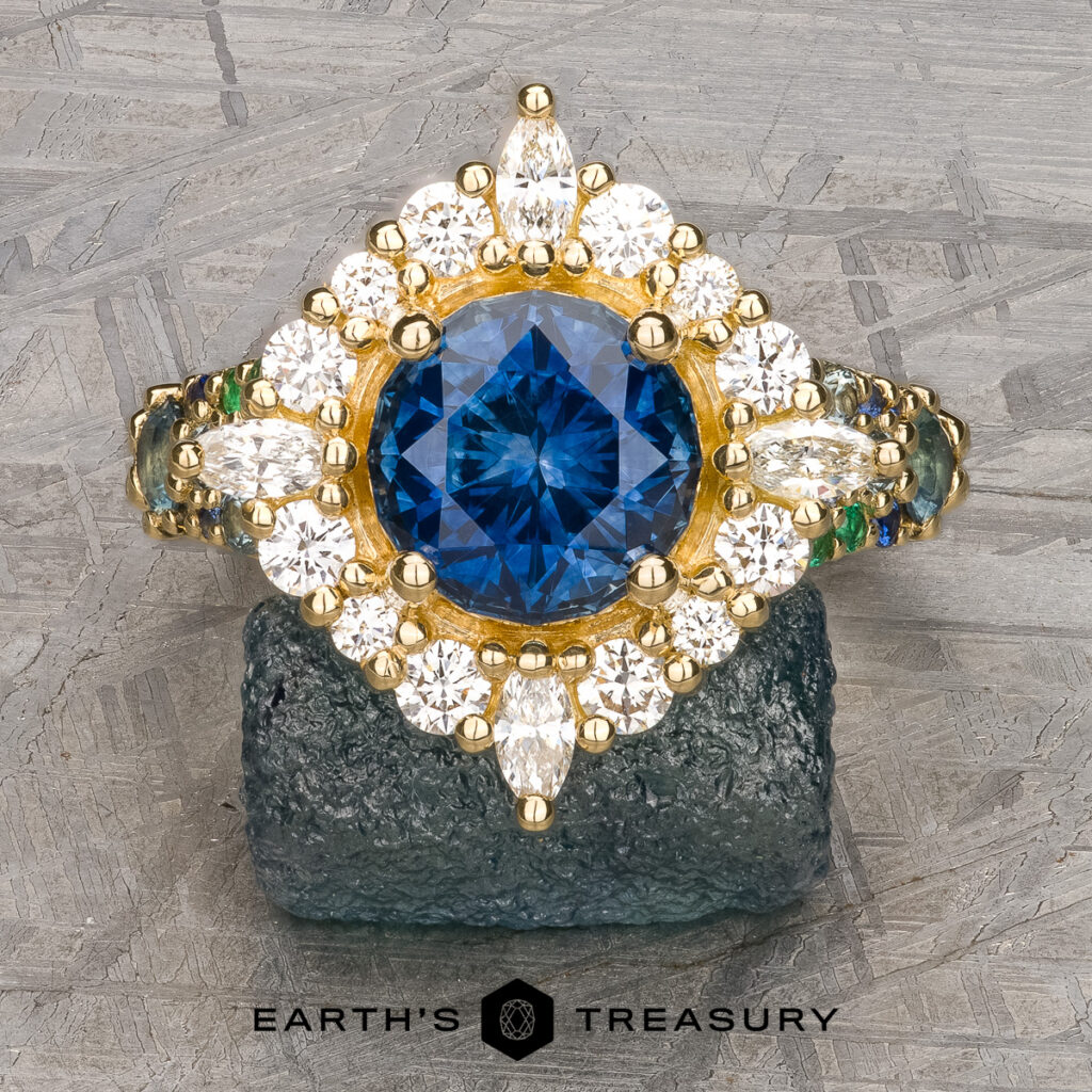 The "donovan, ned and bint-al hussein, raiyah [custom gallery]" gold ring features a large blue gemstone center with white and marquise diamonds, shown upright on a dark stone with the "Earth's Treasury" logo below.