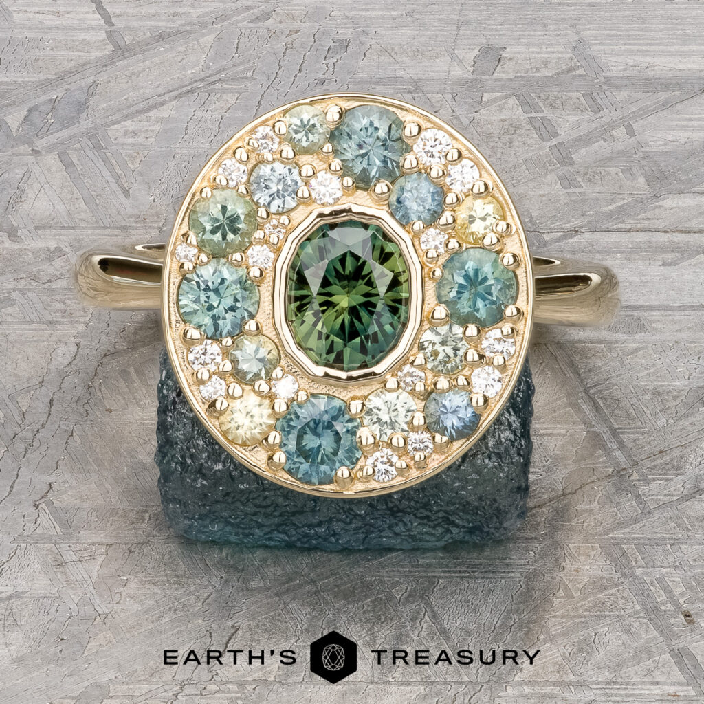A "Whitaker, Wes [custom gallery]" gold ring featuring a round green center stone, accented with blue, green, and white gems, displayed on rough crystal with the "Earth's Treasury" logo below.