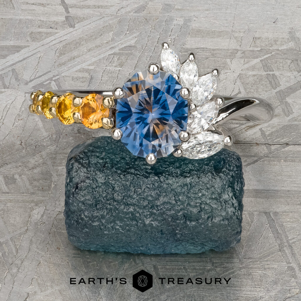 A Kapetanakos, Sherri [custom gallery] silver ring features a round blue gemstone, marquise white stones, and small yellow stones, shown on a rough blue rock.