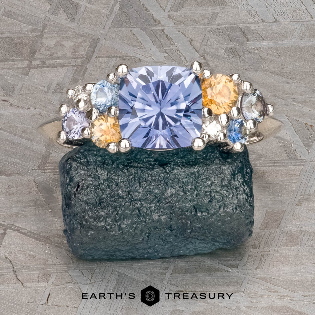 The Harris, Chris and Schuster, Rachel 1 ring features a large blue gemstone set on a textured silver band, displayed on rough crystal with a patterned metal background.