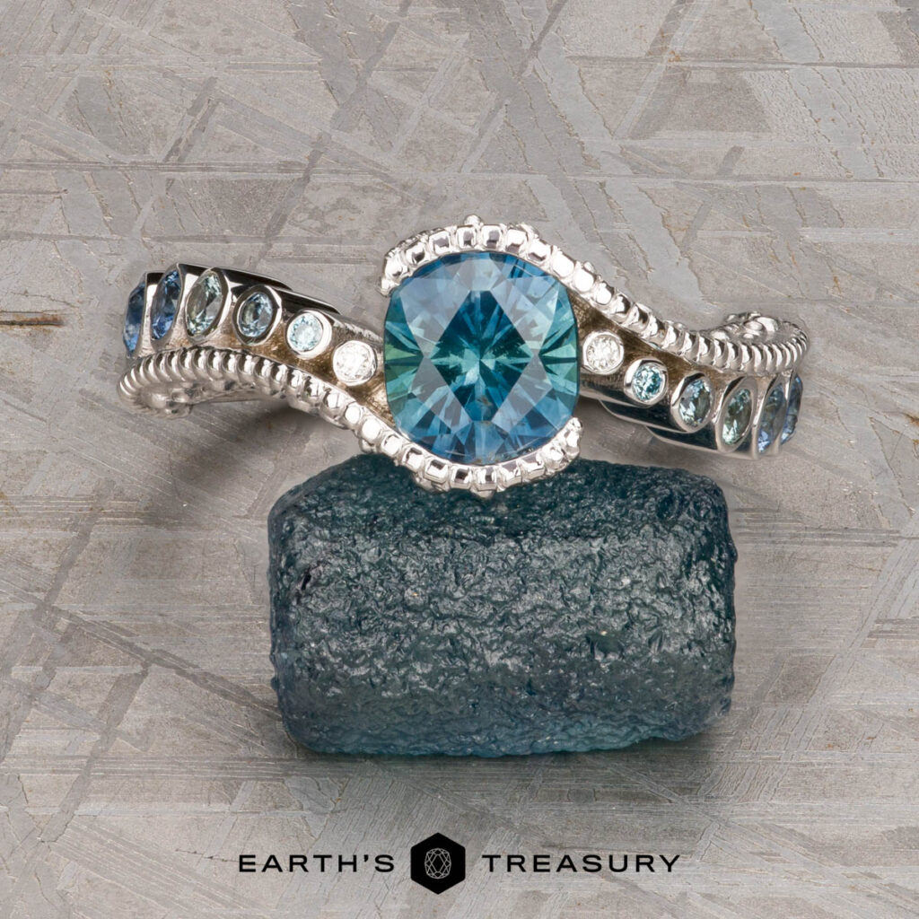 The Simmons, Tim and Carter, Shannon 1 [custom gallery] ring features a large blue gemstone centerpiece with smaller blue stones, displayed on a rough blue crystal above the "Earth's Treasury" logo.