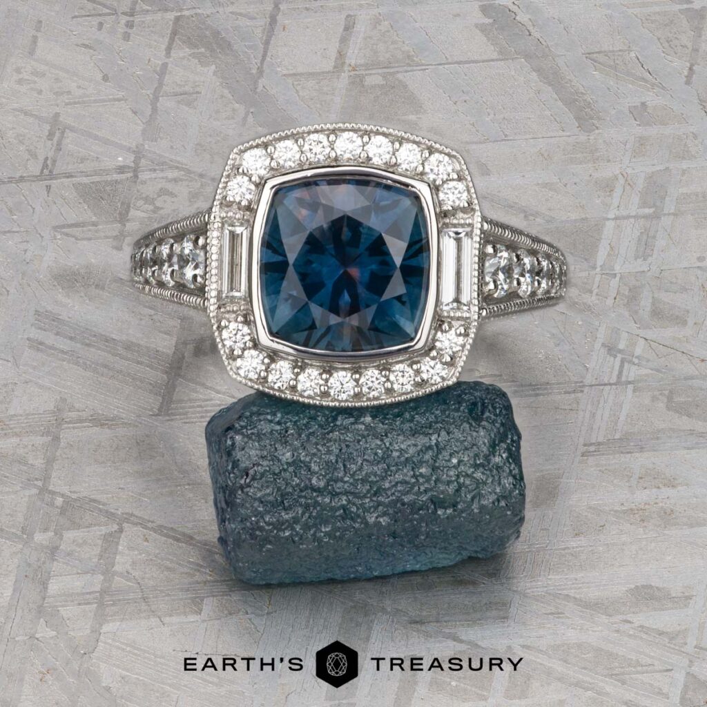 The Remer, Julie and Greenberg, Mark custom gallery showcases a blue gemstone ring with a diamond halo, displayed above a rough blue stone on a textured gray surface with "Earth's Treasury" beneath.