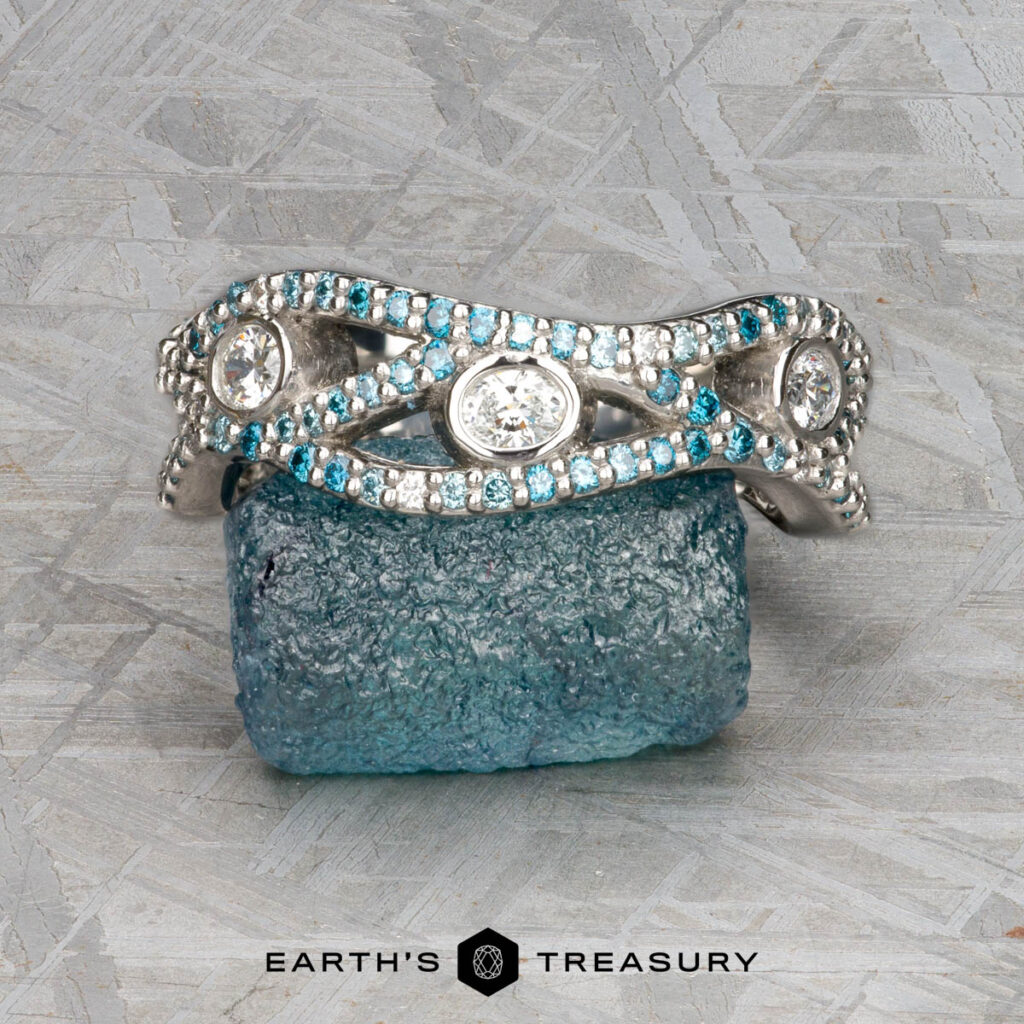 The "Mehta, Tahnee 5 [custom gallery]" silver ring adorned with white and blue gemstones rests on a rough blue stone, set against a textured gray background with the Earth’s Treasury logo at the bottom.