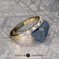 The French-Cut Pave Yogo Band in 18K yellow gold, featuring alternating blue and clear gemstones, lies beside a rough blue stone on a patterned metallic surface. "EARTH'S TREASURY" is displayed at the bottom.