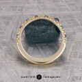 The French-Cut Pave Yogo Band in 18K yellow gold, featuring alternating blue and clear gemstones, lies beside a rough blue stone on a patterned metallic surface. "EARTH'S TREASURY" is displayed at the bottom.