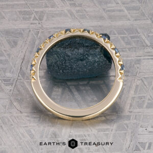 The French-Cut Pave Yogo Band in 18K yellow gold, featuring alternating blue and clear gemstones, lies beside a rough blue stone on a patterned metallic surface. "EARTH'S TREASURY" is displayed at the bottom.