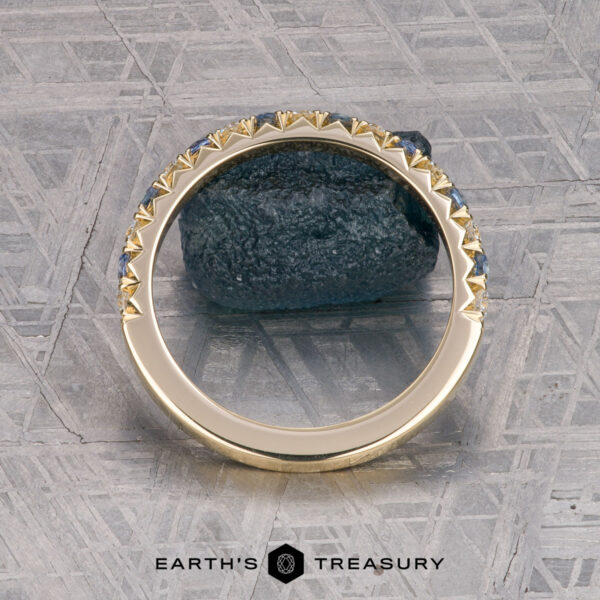 The French-Cut Pave Yogo Band in 18K yellow gold The French-Cut Pave Yogo Band in 18K yellow gold, featuring alternating blue and clear gemstones, lies beside a rough blue stone on a patterned metallic surface. "EARTH'S TREASURY" is displayed at the bottom.