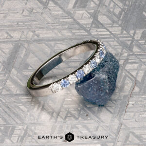A platinum Hand-Cut Pave Yogo Band ring with a jagged, mountain-inspired top edge stands upright before a dark stone on a geometric surface. "Earth's Treasury" appears at the bottom of the image.