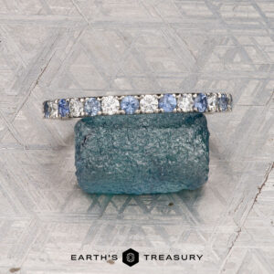 A platinum Hand-Cut Pave Yogo Band ring with a jagged, mountain-inspired top edge stands upright before a dark stone on a geometric surface. "Earth's Treasury" appears at the bottom of the image.