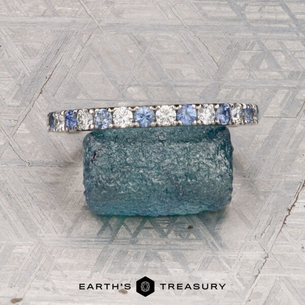 The Hand-Cut Pave Yogo Band in platinum A platinum Hand-Cut Pave Yogo Band ring with a jagged, mountain-inspired top edge stands upright before a dark stone on a geometric surface. "Earth's Treasury" appears at the bottom of the image.