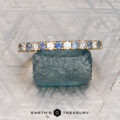 The French-Cut Pave Yogo Band in 18K yellow gold, featuring alternating blue and clear gemstones, lies beside a rough blue stone on a patterned metallic surface. "EARTH'S TREASURY" is displayed at the bottom.
