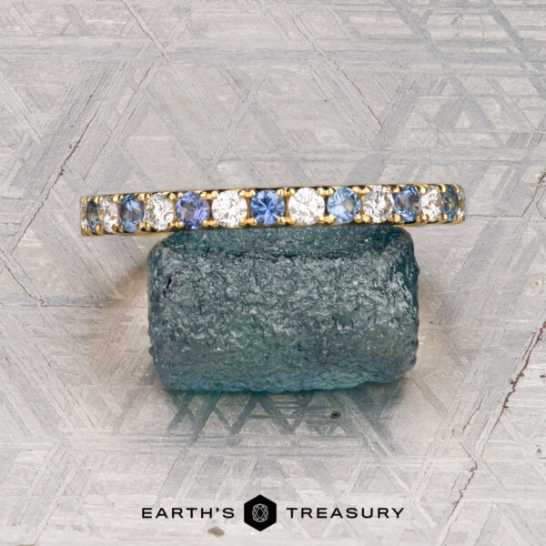 The French-Cut Pave Yogo Band in 18K yellow gold The French-Cut Pave Yogo Band in 18K yellow gold, featuring alternating blue and clear gemstones, lies beside a rough blue stone on a patterned metallic surface. "EARTH'S TREASURY" is displayed at the bottom.