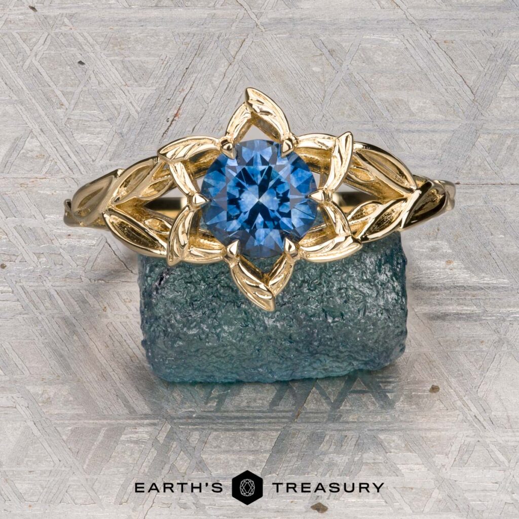 The "Kersh, Scott [custom gallery]" gold ring features a blue gemstone in a floral design, displayed on a textured blue stone background with the "Earth's Treasury" logo below.