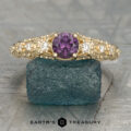 The "Deliah" ring in 14K yellow gold features a 0.56-carat color change garnet center stone and two clear side stones, showcased on a textured greenish-blue rock.