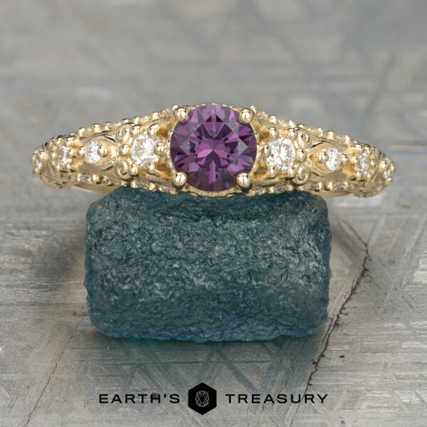 The "Deliah" ring in 14K yellow gold with 0.56-carat color change garnet The "Deliah" ring in 14K yellow gold features a 0.56-carat color change garnet center stone and two clear side stones, showcased on a textured greenish-blue rock.