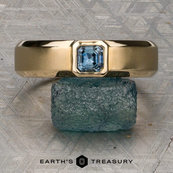 Stacked from 47 images. Method=B (R=7,S=2) A gold ring with an emerald-cut blue gemstone, named "Stacked from 47 images. Method=B (R=7,S=2)," is displayed balanced atop a raw blue stone against a textured metallic backdrop. Crafted in 14K yellow gold and Ready to Ship, the Earth’s Treasury logo appears at the bottom.