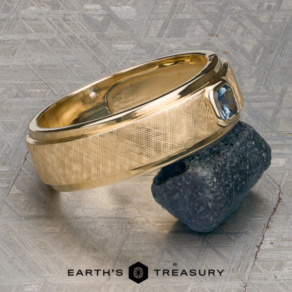Stacked from 49 images. Method=B (R=7,S=2) A "Stacked from 49 images. Method=B (R=7,S=2)" ring in 14K yellow gold, featuring a striking blue gemstone, rests on a rough dark mineral sample atop a textured gray surface. The words "Earth's Treasury" appear at the bottom.