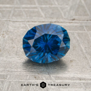 A 1.40-carat heated Montana sapphire, oval-cut and blue, is displayed on a textured gray surface with the text "Earth's Treasury" at the bottom.