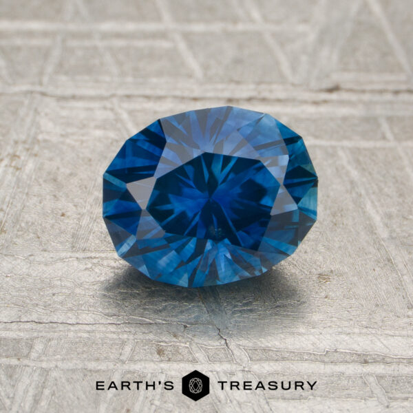 1.40-Carat Rich Blue Montana Sapphire (Heated) A 1.40-carat heated Montana sapphire, oval-cut and blue, is displayed on a textured gray surface with the text "Earth's Treasury" at the bottom.
