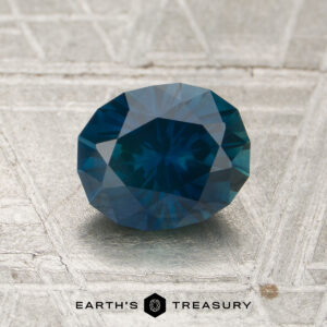 A 1.40-carat oval-cut Montana sapphire (heated) in blue-green rests on a textured metallic surface with the "EARTH'S TREASURY" logo and text below.