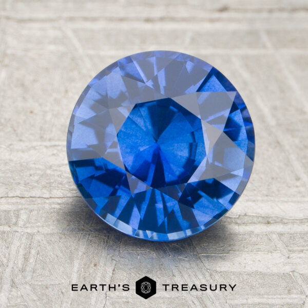 2.49-Carat Sapphire A 2.49-carat round brilliant-cut blue sapphire is showcased on a textured surface, with the "EARTH'S TREASURY" logo and text beneath it.