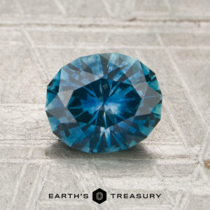 A round, faceted 1.38-carat heated Montana sapphire rests on a textured light gray surface, with "Earth's Treasury" displayed at the bottom.