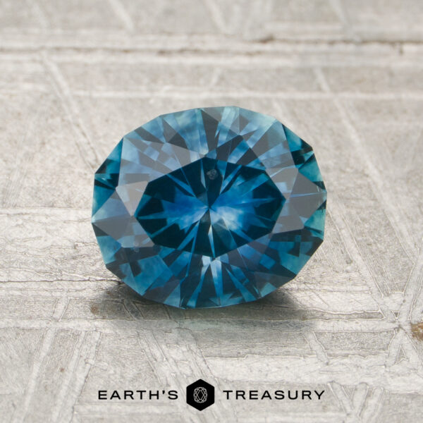 1.38-Carat Teal Blue Montana Sapphire (Heated) A round, faceted 1.38-carat heated Montana sapphire rests on a textured light gray surface, with "Earth's Treasury" displayed at the bottom.