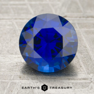 A 2.40-carat round-cut blue sapphire rests on a textured gray surface, with the "Earth’s Treasury" logo and text displayed below.