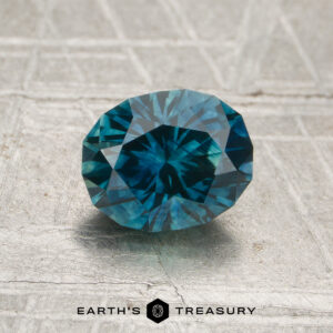 A 1.37-carat heated Montana sapphire, round brilliant-cut and blue, is displayed on a textured metallic surface with the "Earth's Treasury" logo underneath.