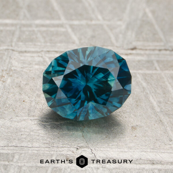 1.37-Carat Rich Teal Montana Sapphire (Heated) A 1.37-carat heated Montana sapphire, round brilliant-cut and blue, is displayed on a textured metallic surface with the "Earth's Treasury" logo underneath.