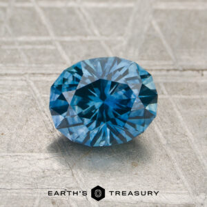 A 1.35-carat heated Montana sapphire with an oval cut sits atop a textured metallic surface, displaying "Earth's Treasury" at the bottom.