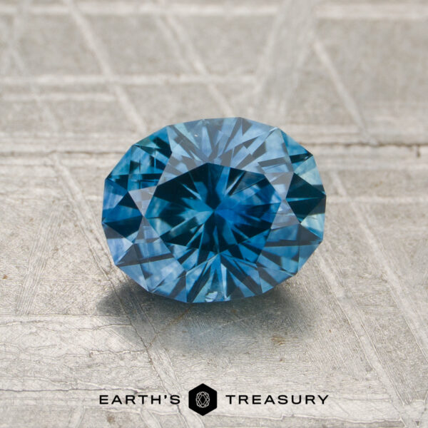 1.35-Carat Teal Blue Montana Sapphire (Heated) A 1.35-carat heated Montana sapphire with an oval cut sits atop a textured metallic surface, displaying "Earth's Treasury" at the bottom.