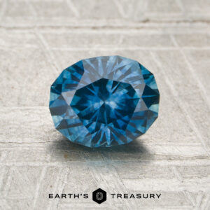A 1.35-carat heated Montana sapphire is displayed on a light textured surface with the EARTH'S TREASURY logo and text below it.
