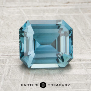 A 2.11-carat emerald-cut blue sapphire is showcased on a light textured surface, with the Earth’s Treasury logo and text visible at the bottom.