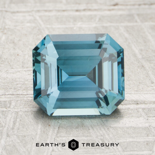 2.11-Carat Sapphire A 2.11-carat emerald-cut blue sapphire is showcased on a light textured surface, with the Earth’s Treasury logo and text visible at the bottom.