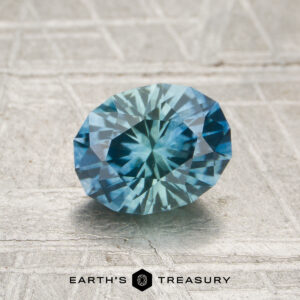 A 1.32-carat heated Montana sapphire, cut in a faceted oval and displaying blue-green hues, rests on a textured light gray surface with the "Earth's Treasury" text at the bottom.