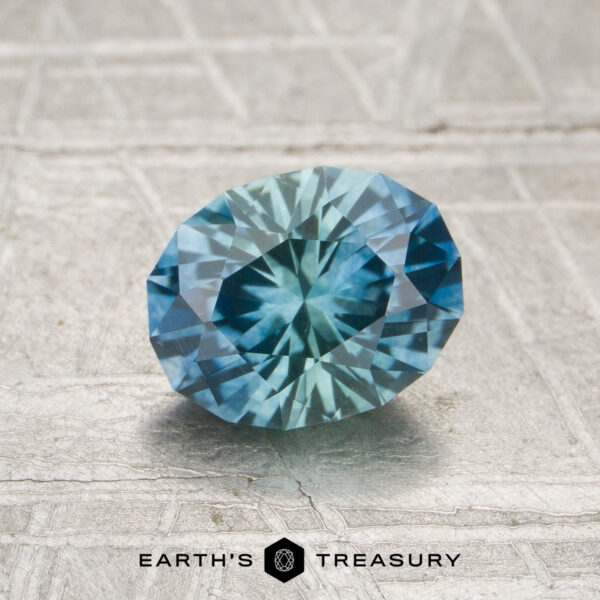 1.32-Carat Teal Blue-Aqua Particolored Montana Sapphire (Heated) A 1.32-carat heated Montana sapphire, cut in a faceted oval and displaying blue-green hues, rests on a textured light gray surface with the "Earth's Treasury" text at the bottom.