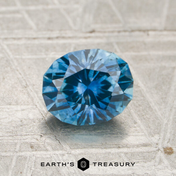 1.31-Carat Teal Blue Montana Sapphire (Heated) A 1.31-carat heated Montana sapphire with a round brilliant cut is displayed on a light textured surface, featuring the "EARTH'S TREASURY" logo and text at the bottom.