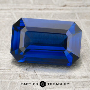 A 2.09-carat, emerald-cut deep blue sapphire is displayed on a textured surface, with the "Earth's Treasury" logo and text at the bottom.