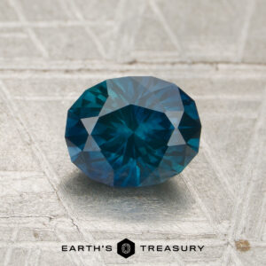 A 1.31-carat heated Montana sapphire, cut in a faceted oval shape, displayed on a light textured surface with "EARTH'S TREASURY" and a logo beneath it.
