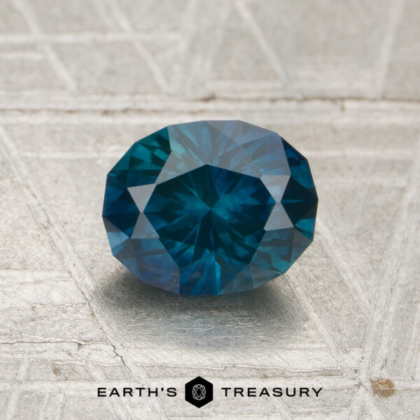 1.31-Carat ich Teal-Peacock Montana Sapphire (Heated) A 1.31-carat heated Montana sapphire, cut in a faceted oval shape, displayed on a light textured surface with "EARTH'S TREASURY" and a logo beneath it.