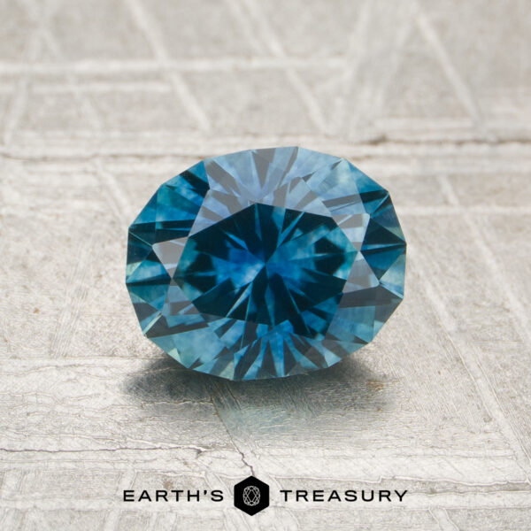 1.31-Carat Teal Blue Montana Sapphire (Heated) A 1.31-carat heated Montana sapphire rests on a textured surface with "EARTH'S TREASURY" and a geometric logo beneath it.