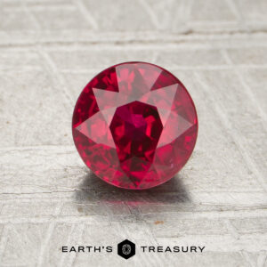 A 1.21-carat round, faceted ruby is displayed on a textured, light background with "EARTH'S TREASURY" written below.