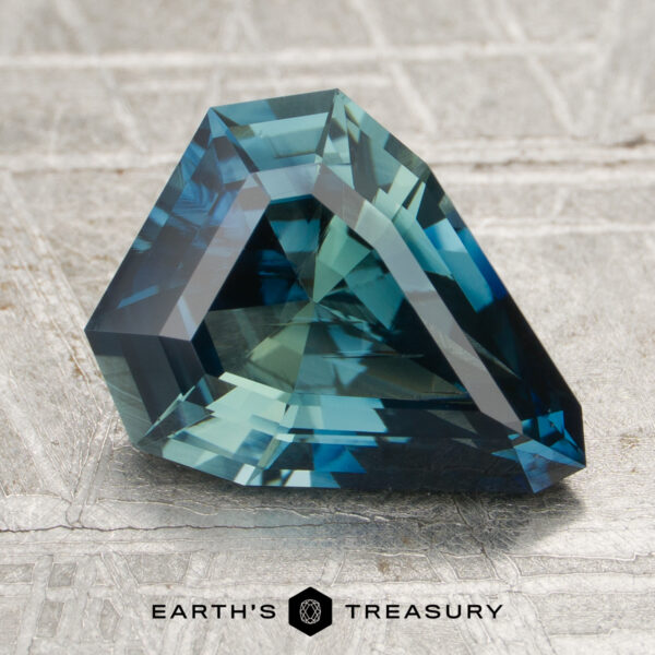3.09-Carat Blue-Green Particolored Madagascar Sapphire (Heated) A 3.09-carat faceted blue-green sapphire with a modern triangular cut rests on a textured gray surface, featuring the "EARTH'S TREASURY" logo at the bottom.