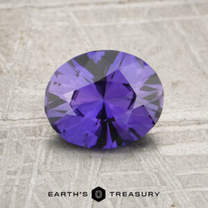 A 1.46-carat unheated Ceylon sapphire with an oval cut is displayed on a textured gray surface, featuring the "Earth's Treasury" logo below.
