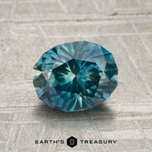 A 1.74-carat heated Montana sapphire with an oval blue-green facet is displayed on a textured light surface, with the Earth's Treasury logo below.