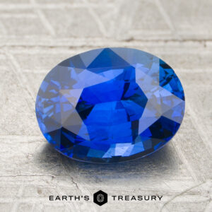 A 2.02-carat sapphire gemstone rests on a textured metallic surface, with "EARTH'S TREASURY" displayed below.