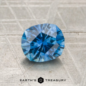 A 1.29-carat heated Montana sapphire is displayed on a textured grey surface with the "EARTH'S TREASURY" logo and text at the bottom.