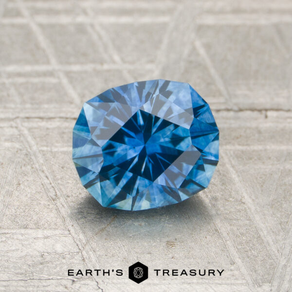 A 1.29-carat heated Montana sapphire is displayed on a textured grey surface with the "EARTH'S TREASURY" logo and text at the bottom.
