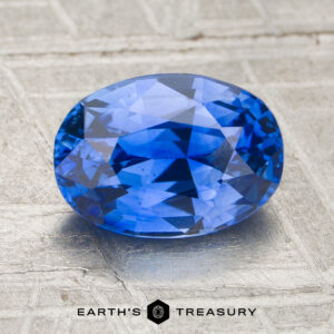 A 1.66-carat sapphire, faceted and oval-shaped, sits on a textured gray surface with "EARTH'S TREASURY" displayed beneath.